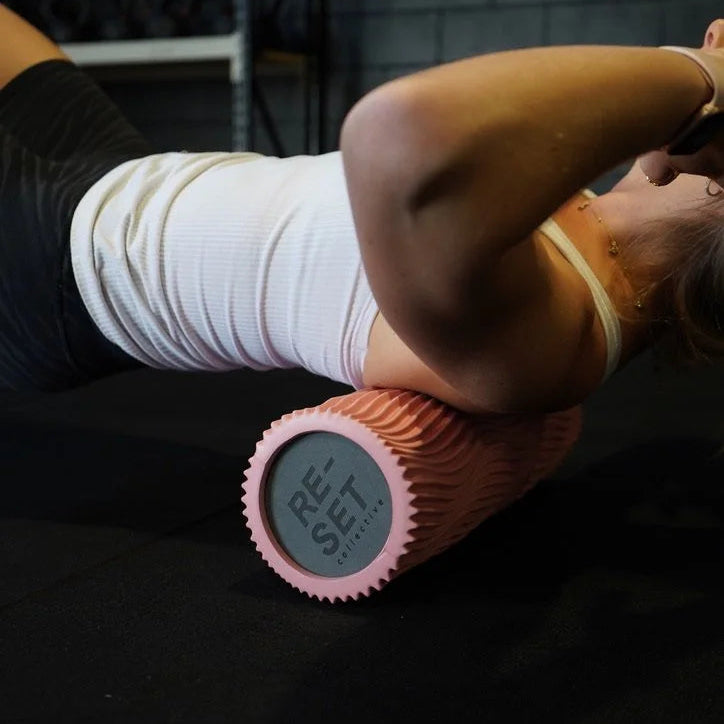 A person lying on a black gym floor uses a pink textured foam roller with a digital display showing 'GET fit'.