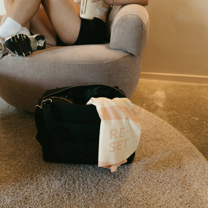 A black duffel bag sits on a carpeted floor in front of a gray lounge chair, with a person reclining in the chair holding a drink. A white towel draped over the bag has pink text reading 'RESET'.