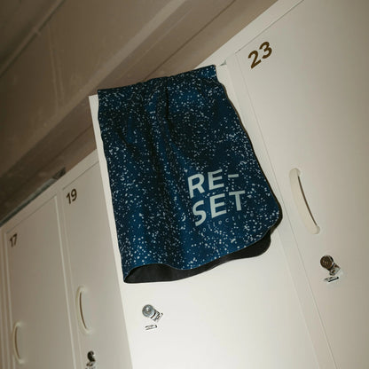 dark blue athletic towel with a white speckled pattern and the word 'RESET' printed on the front, hanging on a white locker in a gym hallway.