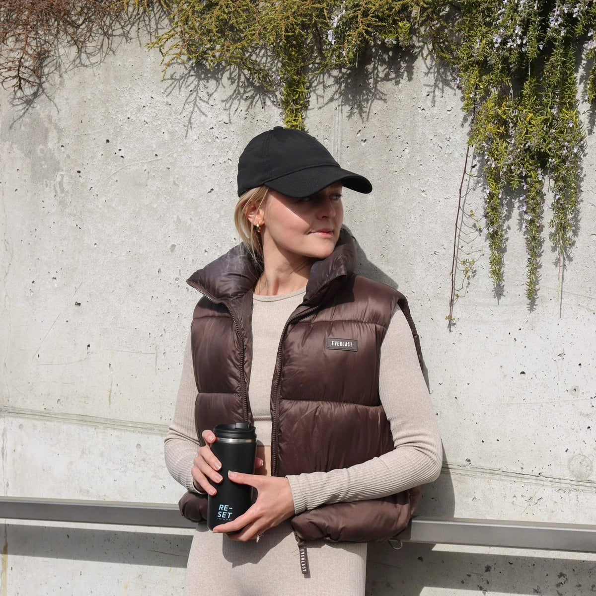 A woman wearing a dark brown puffer vest over a beige long-sleeve top, a black baseball cap, and holding a black travel mug, standing against a textured concrete wall with green trailing plants.