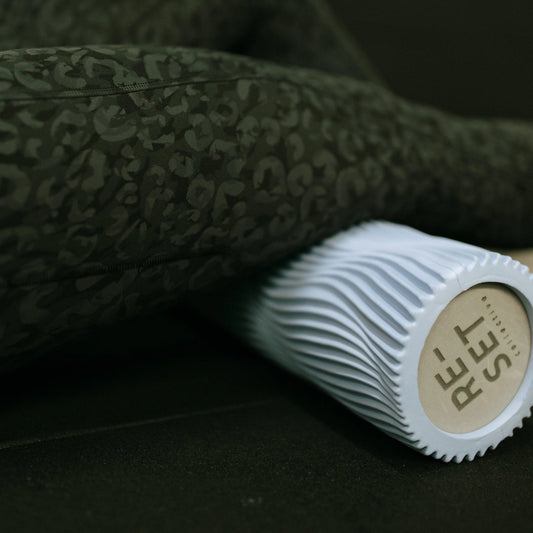 A close-up lifestyle shot of a person's legs resting on a dark textured floor, with a white ribbed foam roller featuring a beige circular logo labeled 'REST' placed in the foreground.